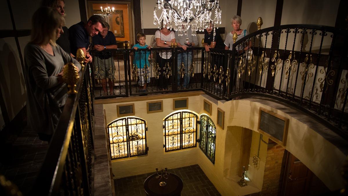 En gruppe mennesker står på en balkon og ser ned mod en smuk foyer med glasmosaikvinduer og en lysekrone.