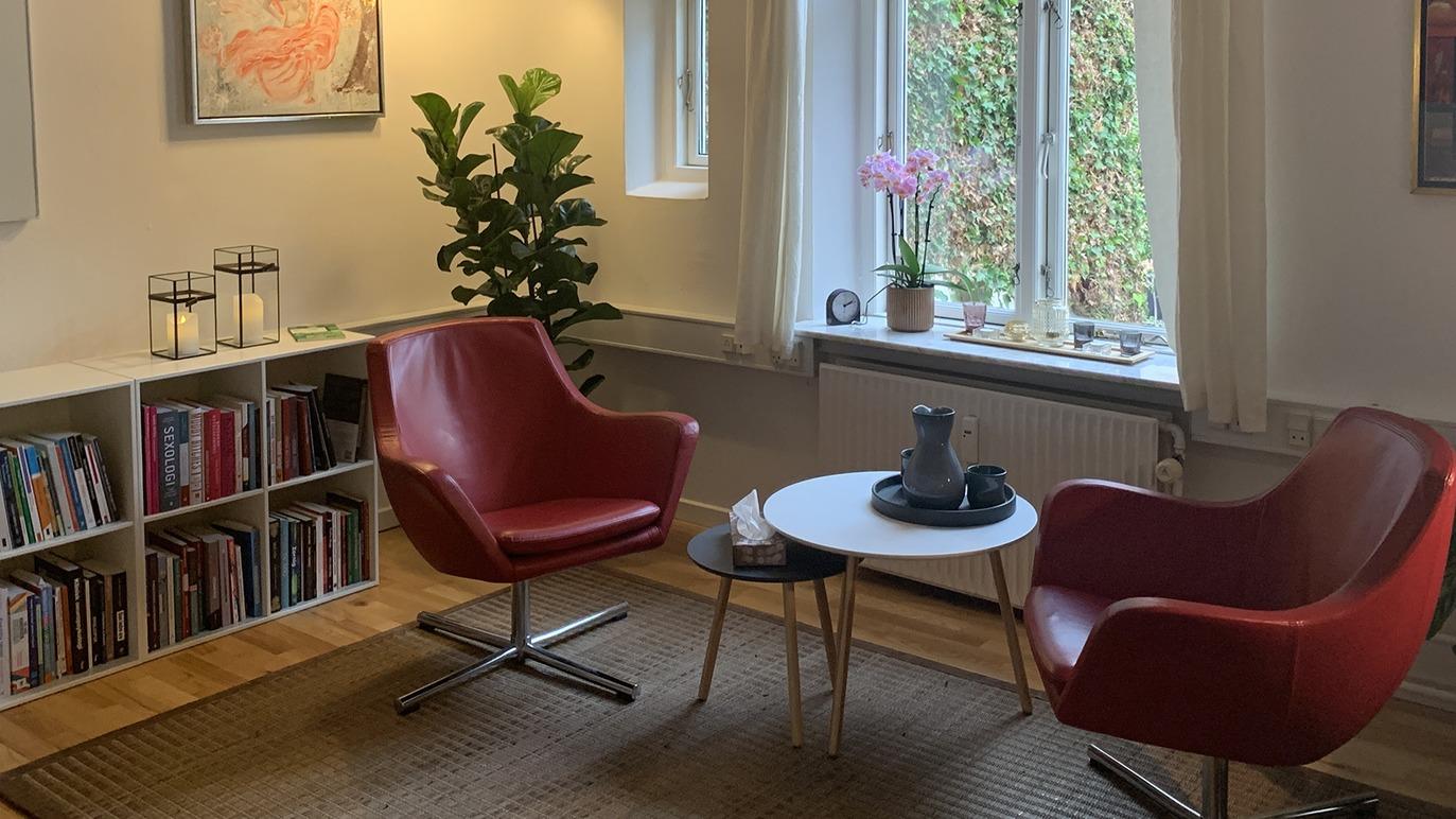 Stue med to røde lænestole, et rundt bord, en plante og bøger i reolen ved vinduet.