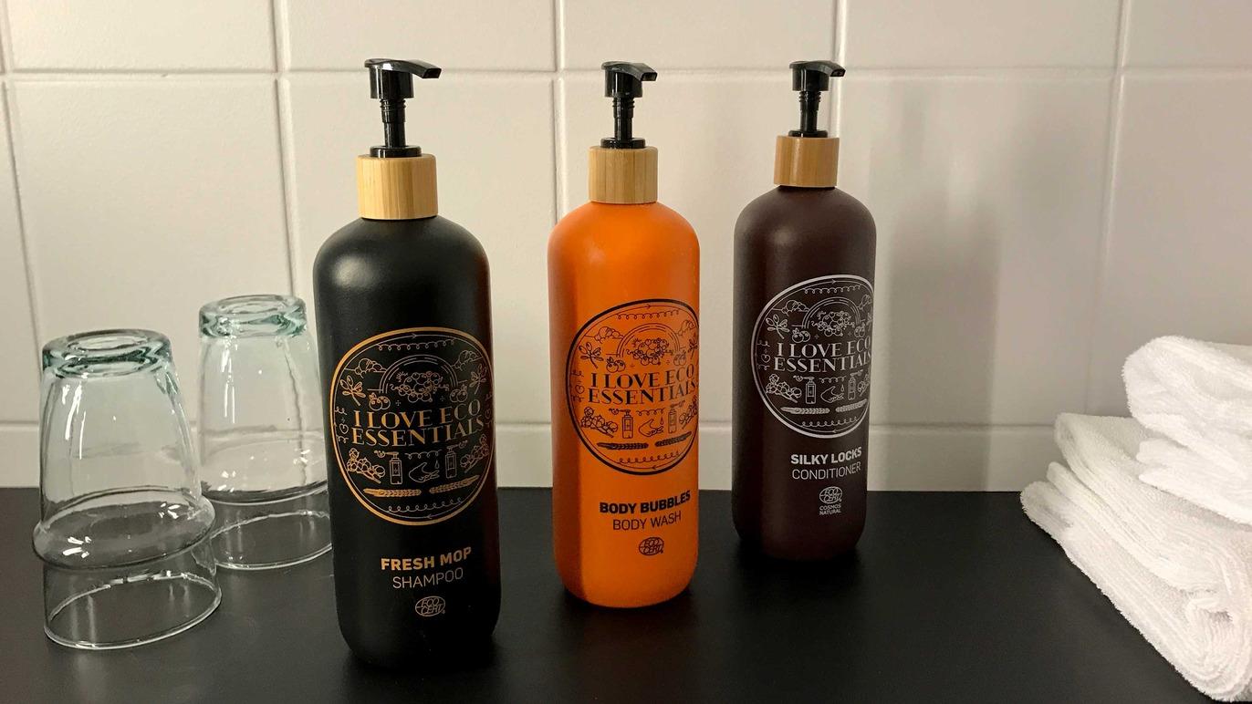 Tre flasker med hårprodukter i forskellige farver står på en sort overflade ved siden af glas og håndklæder.