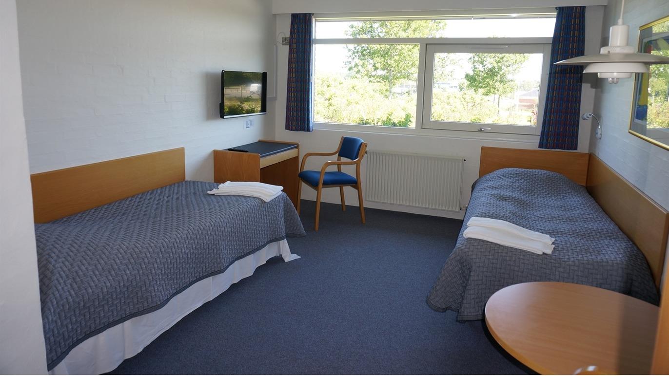 Lyst hotelværelse med to enkeltsenge, tv, skrivebord og stol ved vinduet med udsigt til naturen.