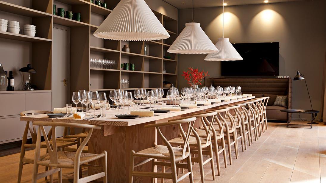 Moderne spisestue med lang træbord, elegante stole og store lamper, dekoreret med glas og blomster.