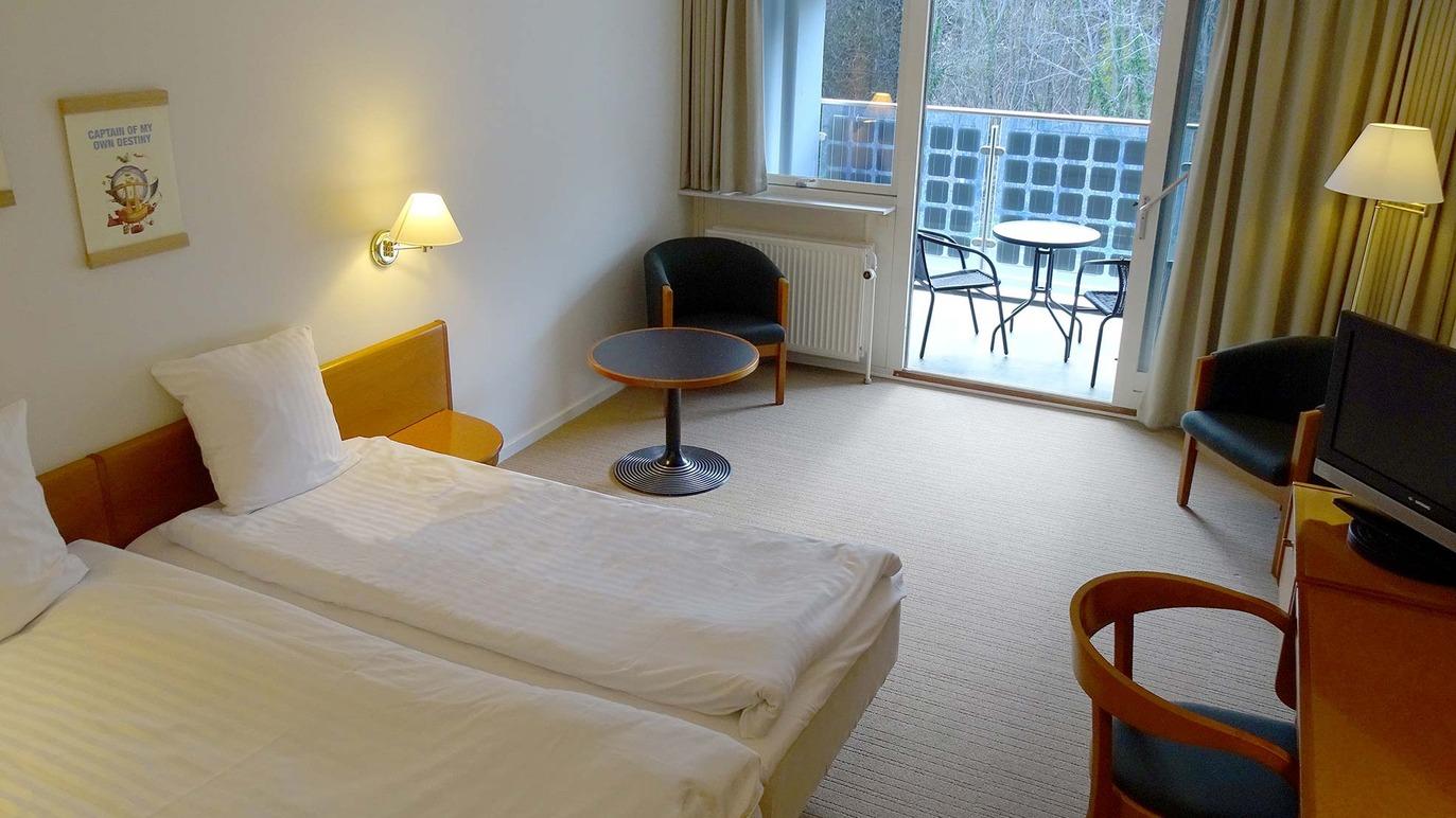 Lyst hotelværelse med to enkeltsenge, skrivebord, stol og balkon med udsigt til haven.