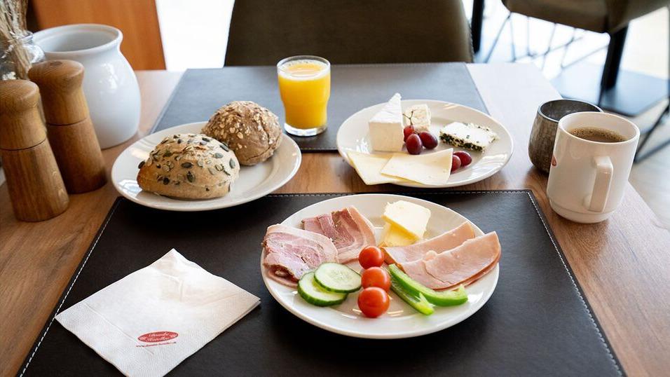 En morgenmadstallerken med skiver af skinke, agurk, cherrytomater og smør, samt brød og juice på bordet.