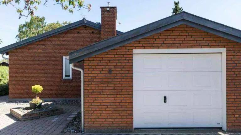 En murstensvilla med garage og indkørsel, omgivet af grønne planter og træer.