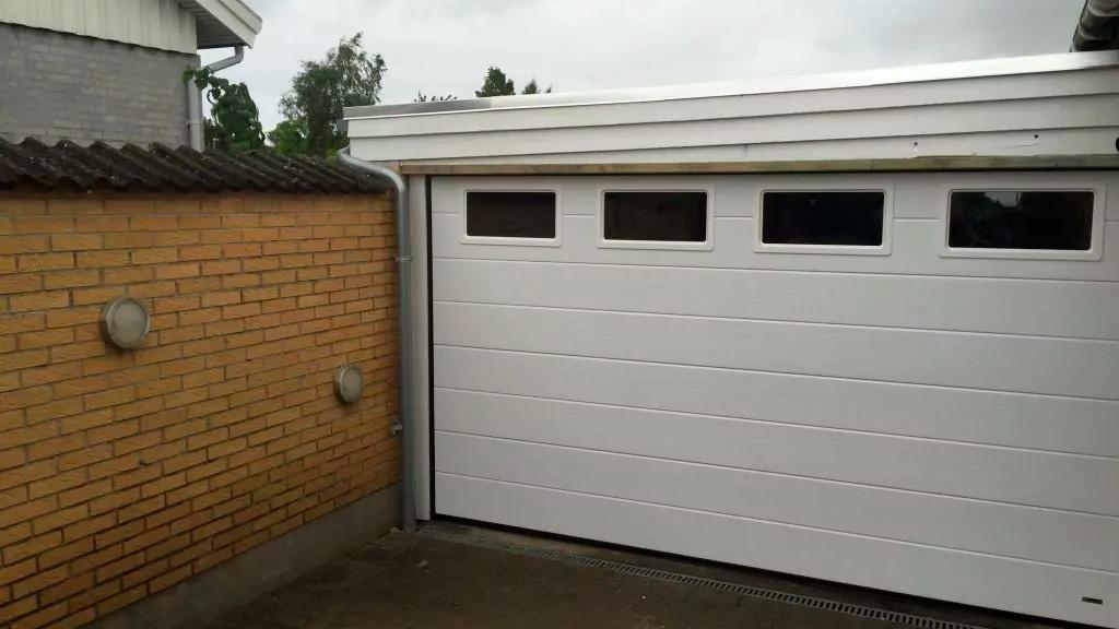 Hvid garageport med vinduer ved en murstensvæg i en boligområde under en grå himmel.