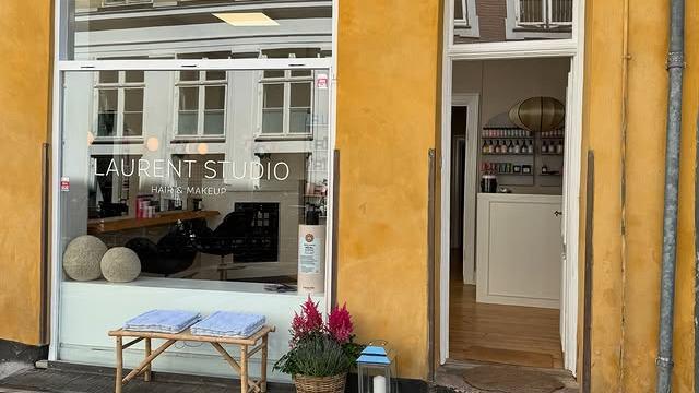En frisørsalon med navnet "Laurent Studio" og en indgang med planter og blomster foran vinduet.