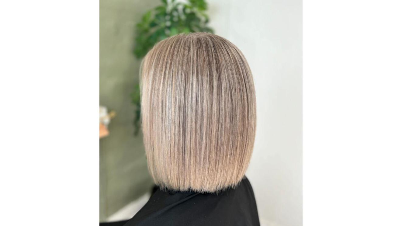 En kvinde med en moderne, lige bob-frisure i lys blond farve, set bagfra med grønne planter i baggrunden.