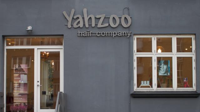 Yahzoo frisørsalon med grå væg og vinduer, der viser frisørprodukter og en plakat af en model.