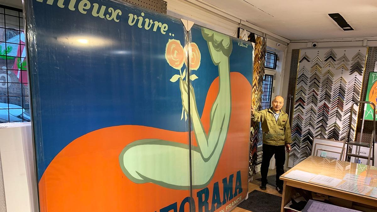 En person står ved siden af en stor vintage plakat med en kvinde, der holder roser, i en butik.