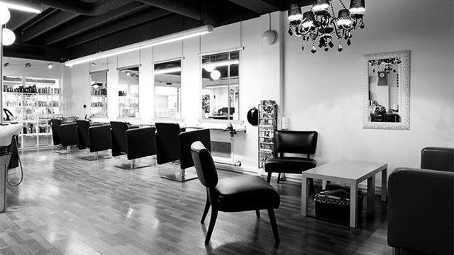 En moderne frisørsalon med sorte stole, spejl og en elegant lysekrone i et lyst rum.