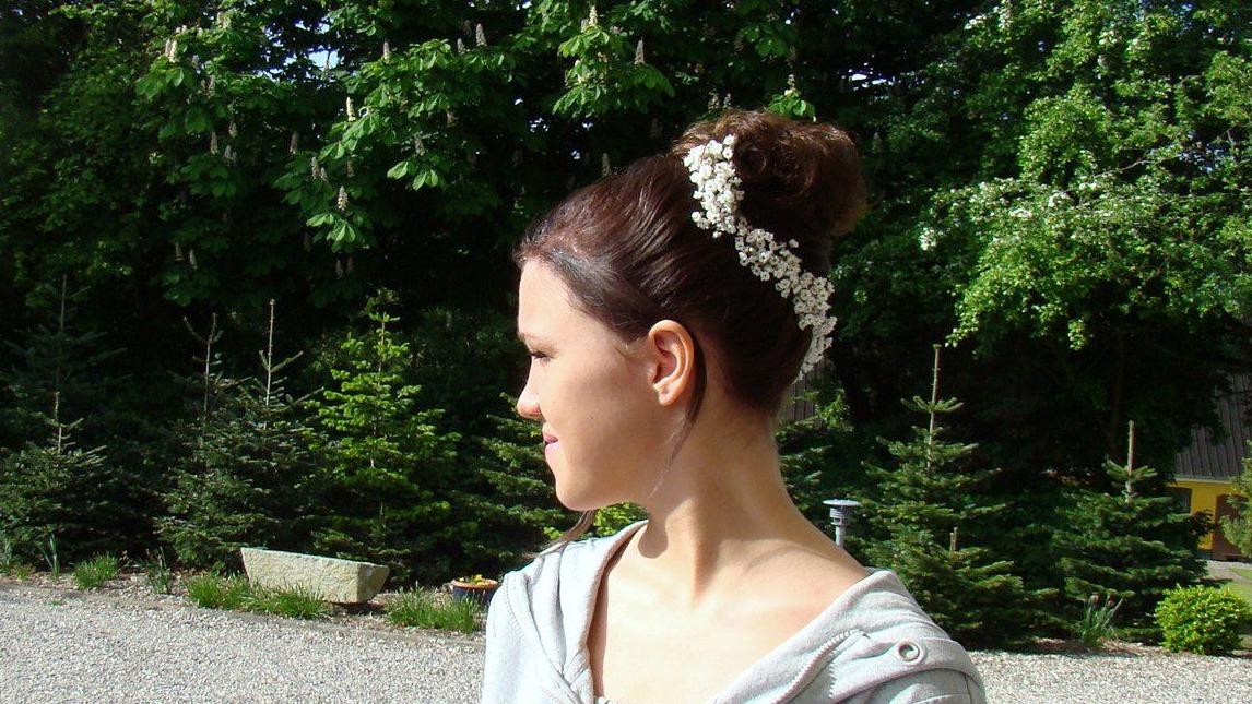 Ung kvinde med en elegant opsat frisure og blomster i håret, står foran en grøn baggrund med træer.