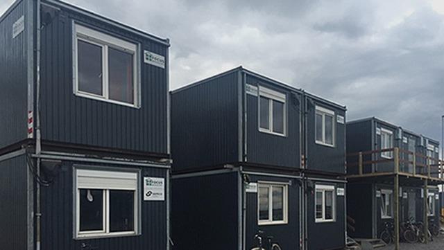 Grå containere stablet i en række under en overskyet himmel.