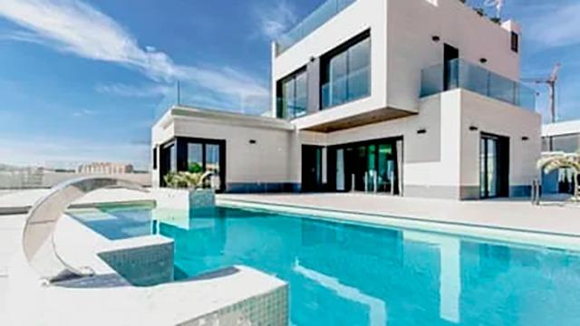 Moderne villa med swimmingpool og glasfacade under en klar blå himmel.