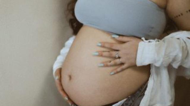 Gravid kvinde i grå top holder om sin mave med lange negle og smykker.