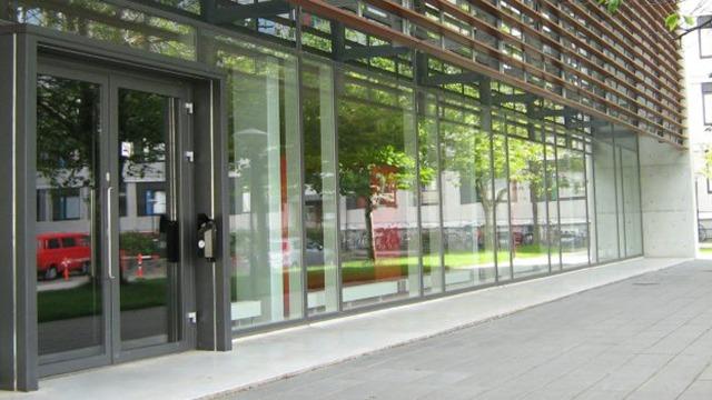 Moderne bygning med glasfacade og dør, omgivet af grønne træer og en græsplæne.
