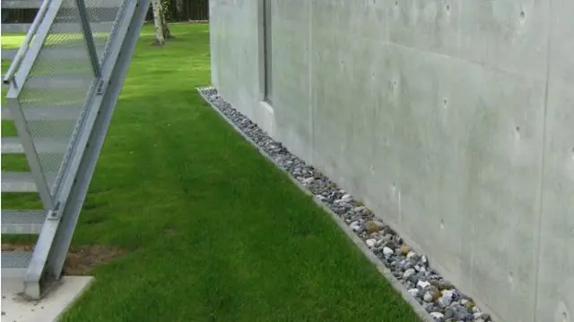 Græsplæne ved en betonvæg med småsten langs kanten og en metaltrappe i baggrunden.