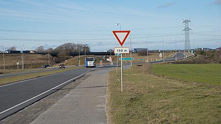 Vejskilt med vigepligt og bus på vej mod kryds, med grønne marker i baggrunden.