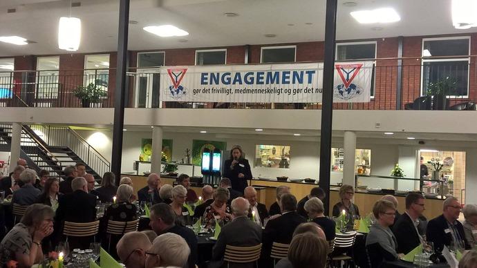 En festlig middag med gæster, der sidder ved borde, og en banner med teksten "ENGAGEMENT" i baggrunden.