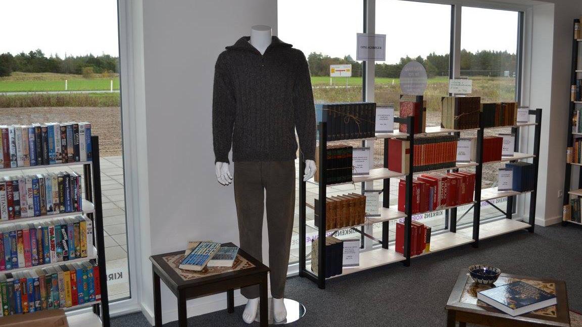 En mannequin iført en grå sweater og bukser står i en boghandel med bøger på hylderne.