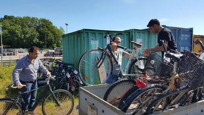 Mænd læsser cykler i en trailer ved en genbrugsplads, omgivet af containere og grønt område.