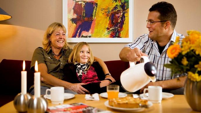 Familie sidder sammen i stuen, nyder te og kage, med blomster og kunstværk i baggrunden.
