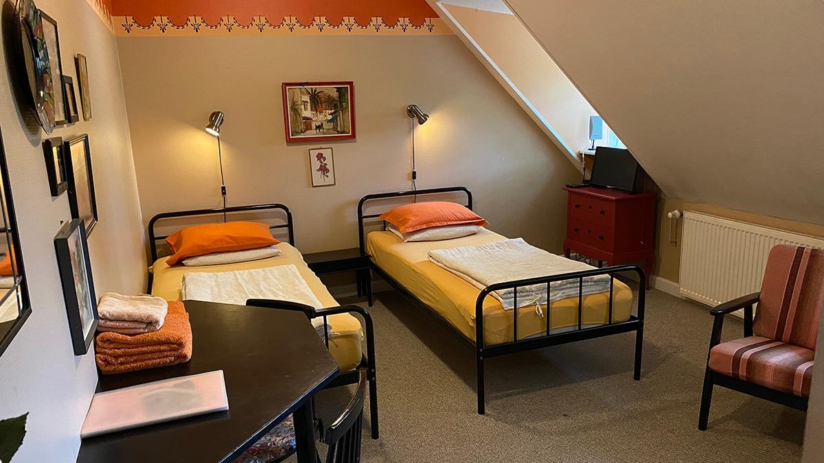 Lyst værelse med to enkeltsenge, skrivebord, stol og dekorative billeder på væggene.