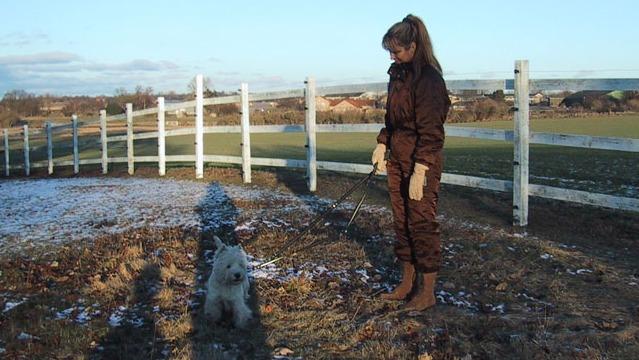 En kvinde i brunt tøj står på en snefyldt mark med en lille hund i snor.