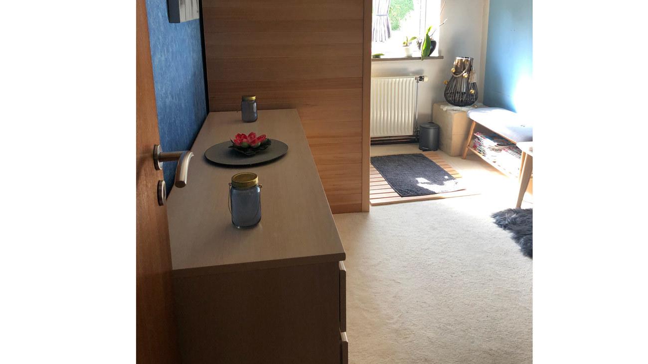 Indgang til et lyst rum med en trækommode, en vase med blomster og en sofa ved vinduet.