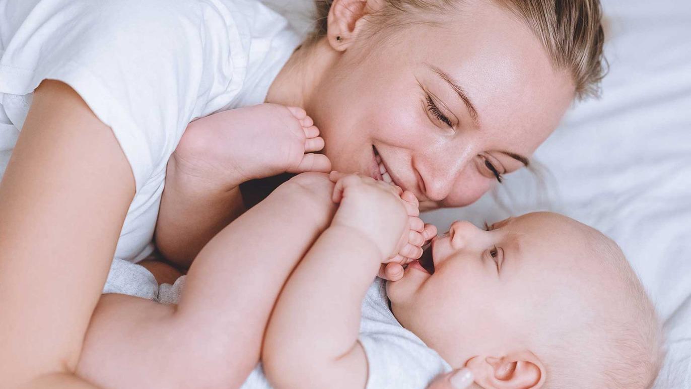 Mor og baby leger sammen i sengen, smilende og holder hinandens hænder.