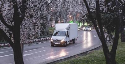 En hvid varevogn kører på en vej omgivet af blomstrende træer i en bypark.