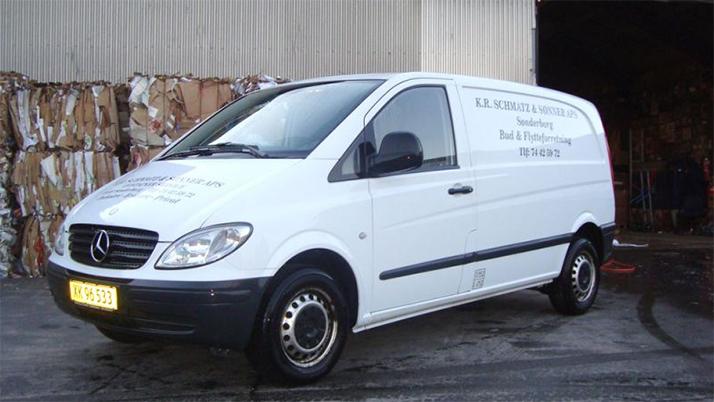 Hvid varevogn fra Mercedes med tekst på siden, parkeret foran en lagerbygning.