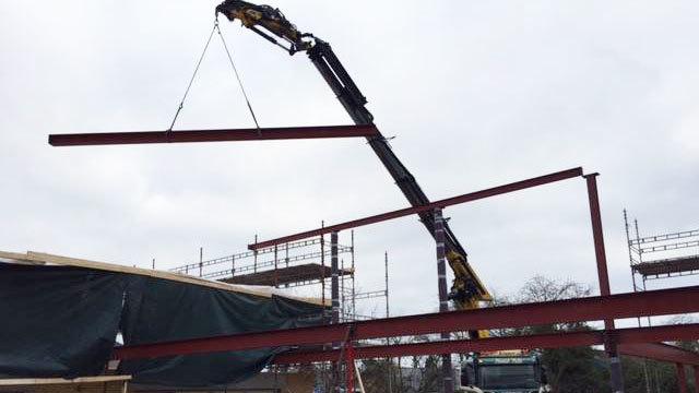 Kran løfter en stålbjælke over en byggeplads med stilladser og bygningselementer i baggrunden.