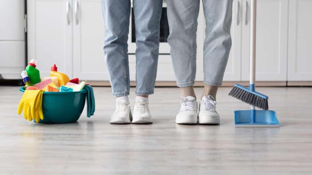 To personer i sneakers står på et rengjort køkkengulv med en spand og rengøringsredskaber ved siden af.
