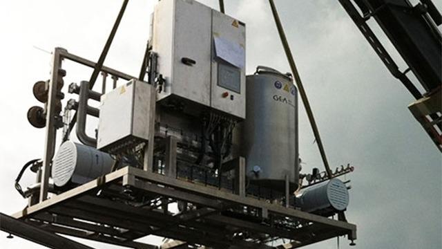 En stor kran løfter en industriel maskine med motorer og kontrolpanel under en skyet himmel.