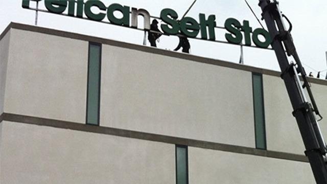 Arbejdsfolk hænger et skilt op med teksten "Pelican Self Storage" på en bygning.
