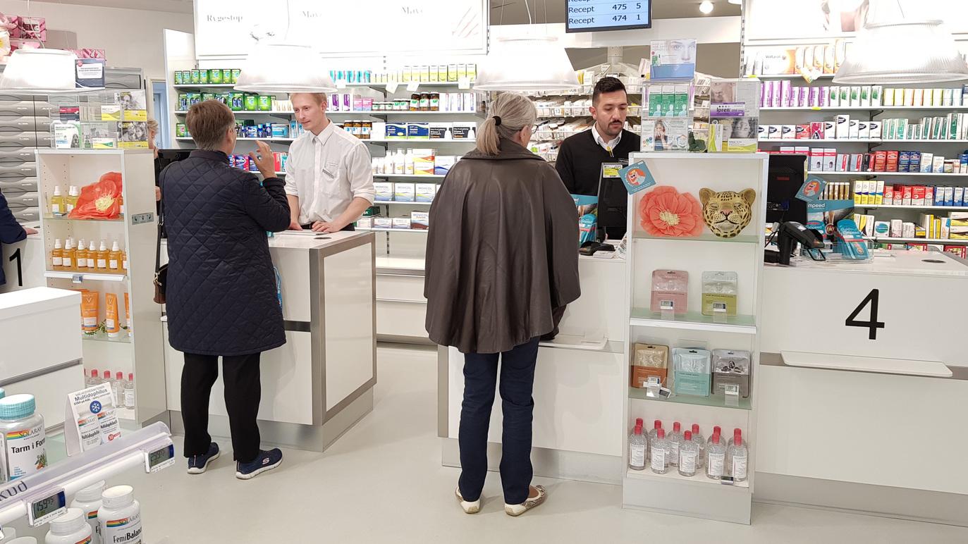 Kunder i en apotekbutik, der interagerer med personalet ved disken og ser på produkter på hylderne.