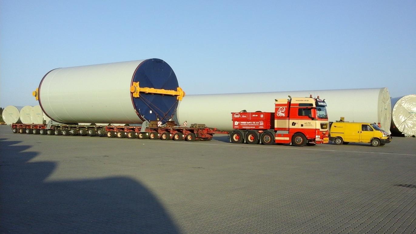 En stor lastbil transporterer en cylindrisk beholder på en trailer i en havn.
