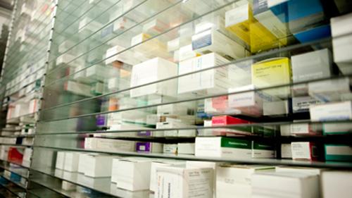En hylde fyldt med medicinæsker i forskellige farver og størrelser, organiseret bag glas.