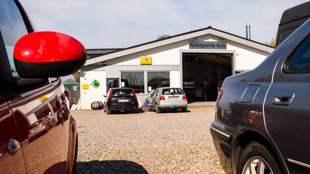 Bilværksted med flere biler parkeret foran og en garageåbning i baggrunden. Solskin og blå himmel.