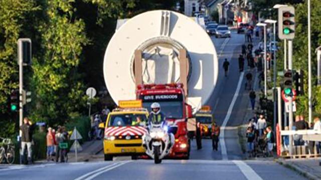 En stor transportvogn med en rund genstand kører gennem byen, eskorteret af politimotorcykler og biler.