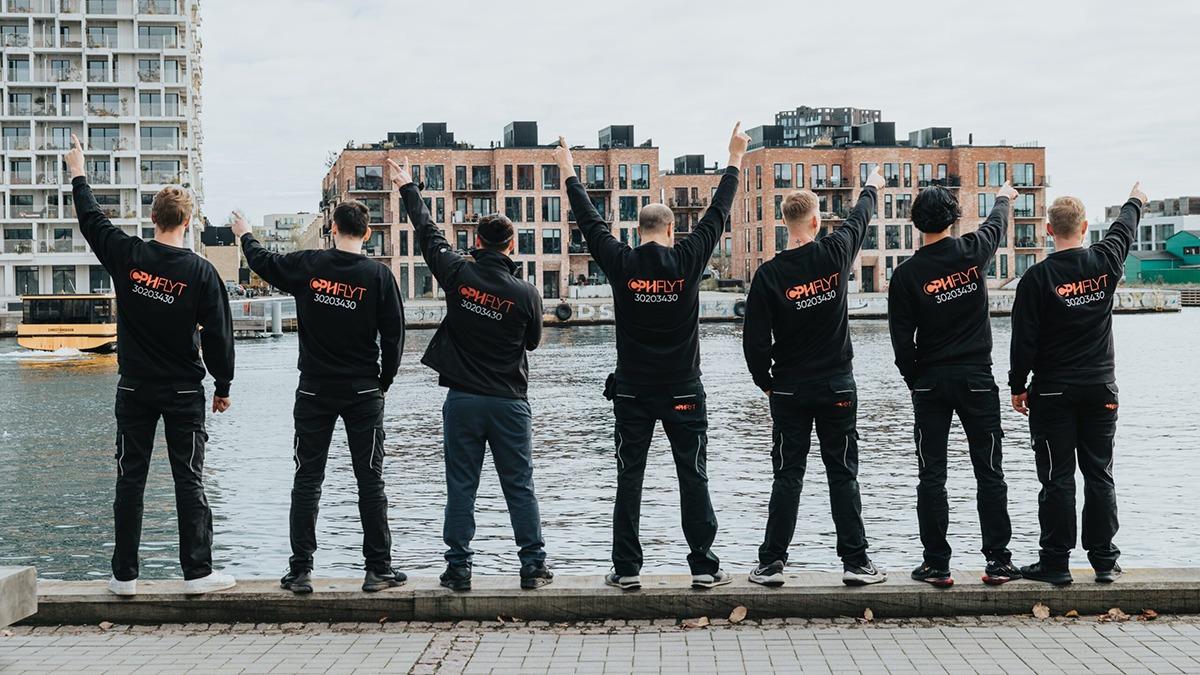 Gruppe af seks personer i sorte sweatshirts, der står ved vandet og vinker mod kameraet i København.