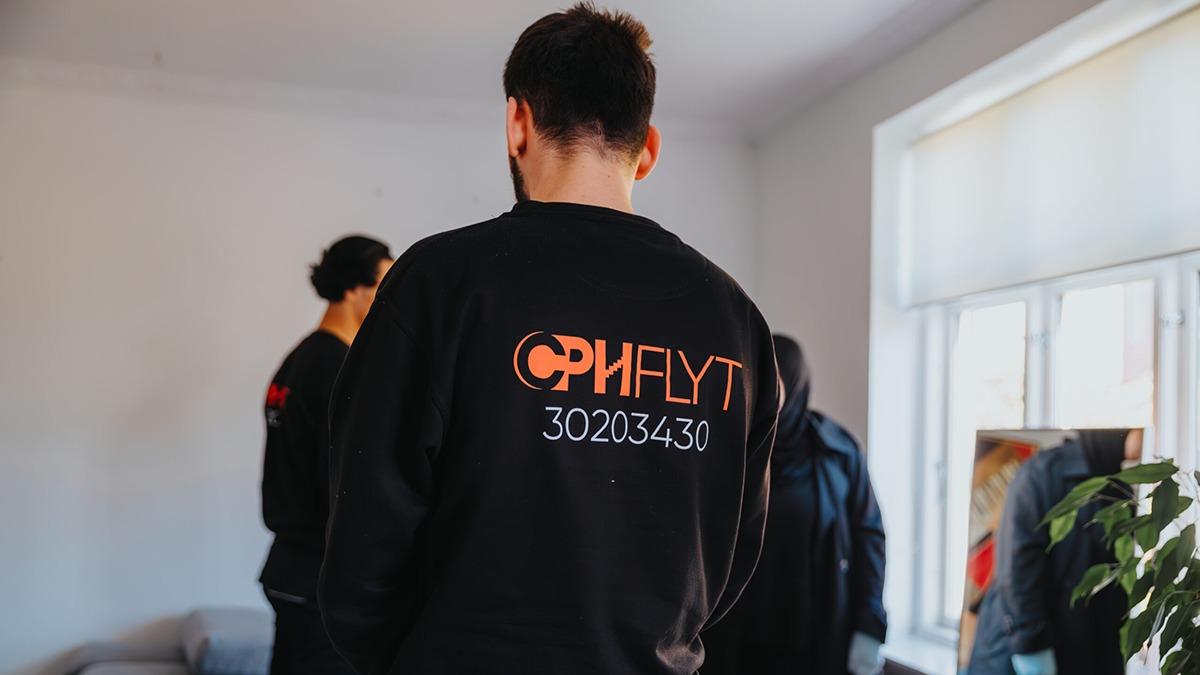 En person iført en sort sweatshirt med teksten "OPHLYT" og et telefonnummer, står i et lyst rum.