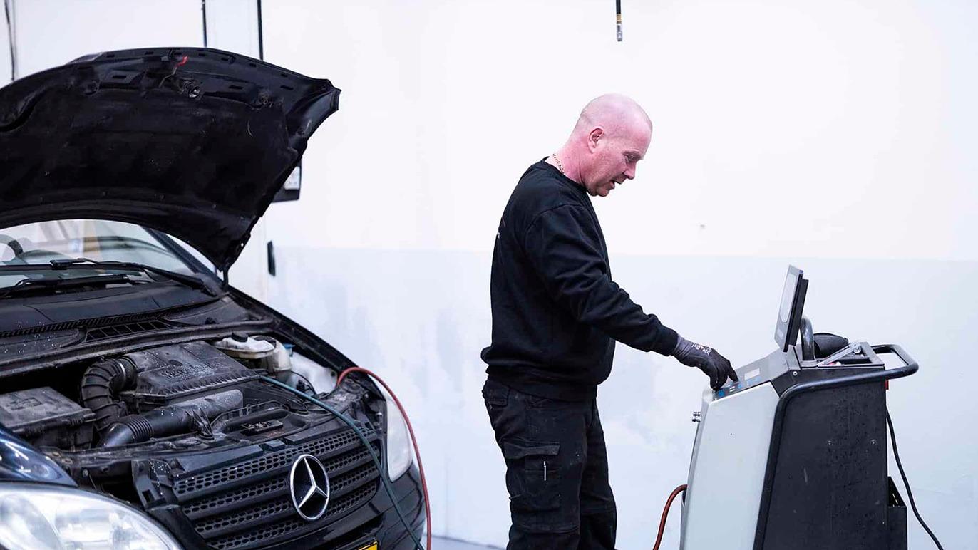 En mekaniker arbejder på en Mercedes bil med åben motorhjelm og diagnosticeringsudstyr i værkstedet.