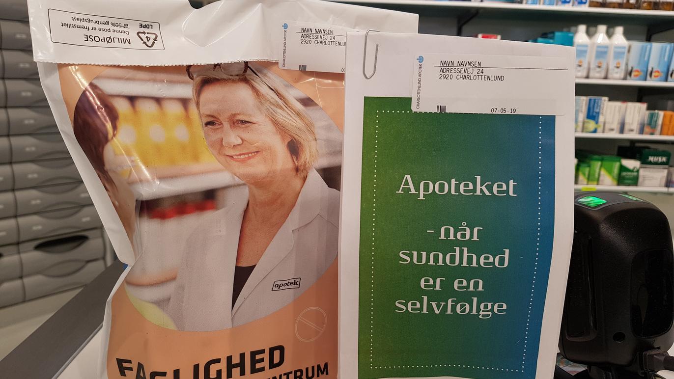 En kvinde smiler ved en apotekvæg med produkter og en plakat om sundhed.