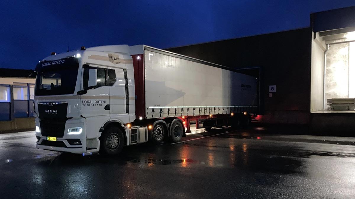 En hvid lastbil parkeret ved en bygning om aftenen, med en lysende facade og våde veje.