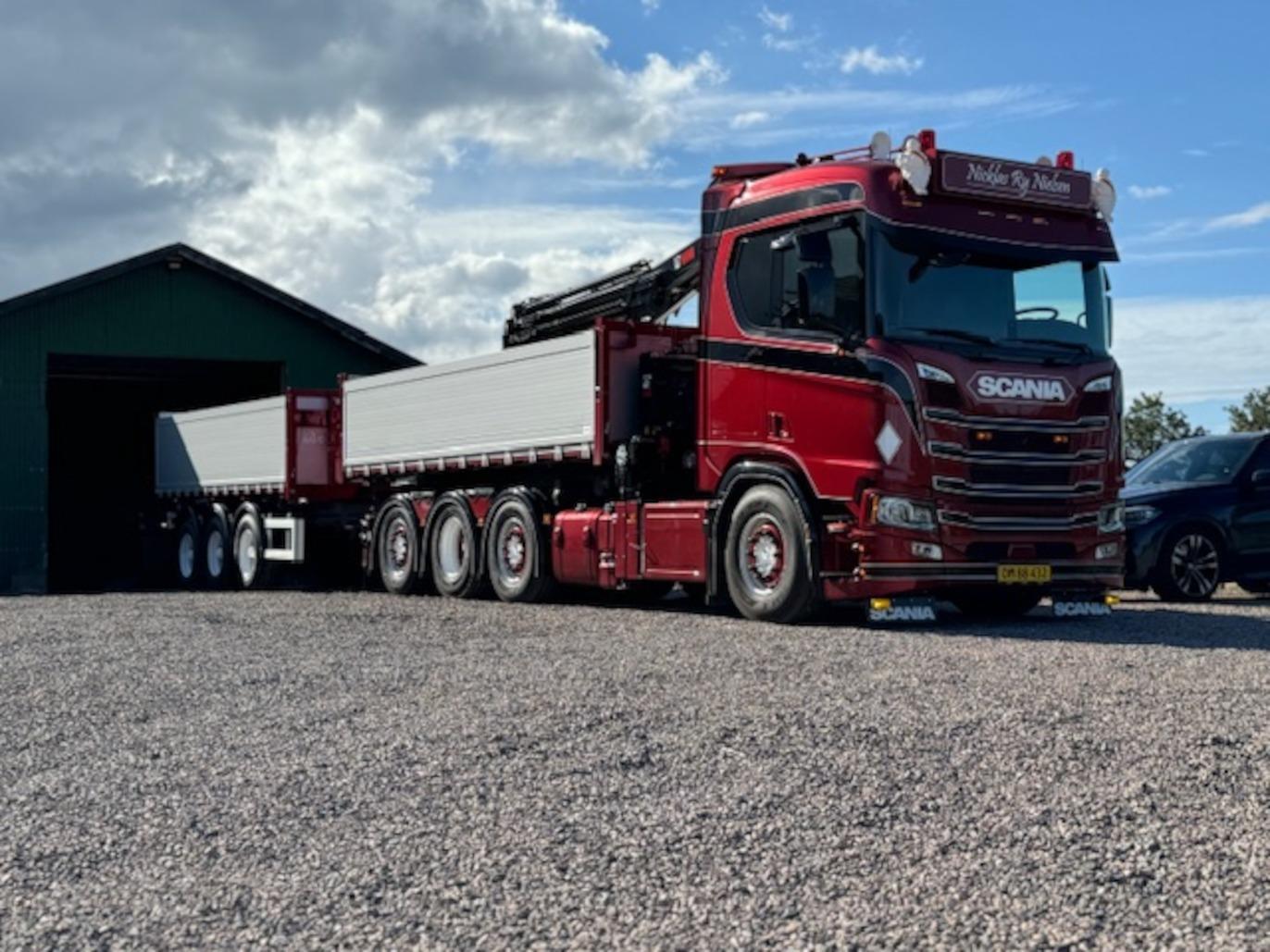 En rød Scania lastbil med en trailer parkeret foran en grøn bygning på en grusvej.