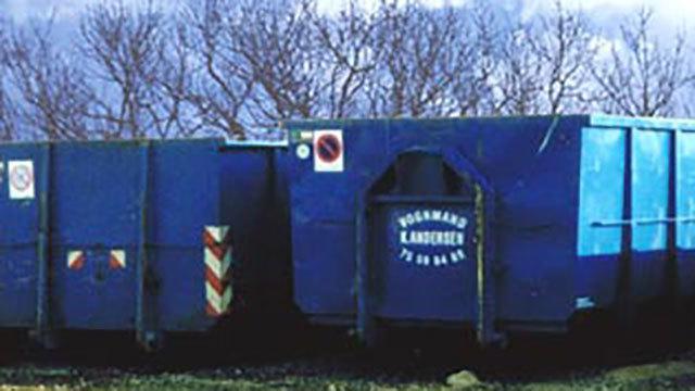 Blå affaldscontainer med tekst og advarselsmærker, placeret i et åbent område med træer i baggrunden.