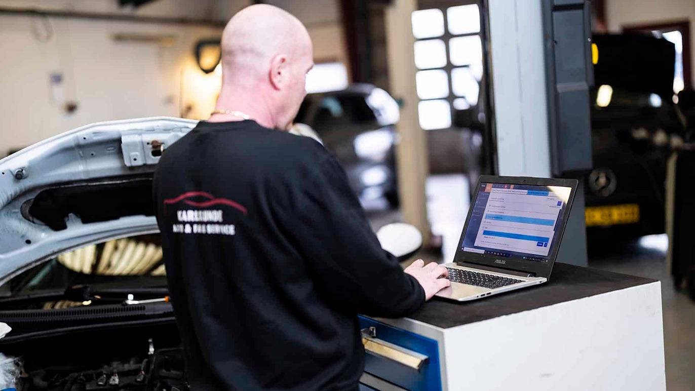Mekaniker arbejder på en bærbar computer i et autoværksted med en åben bilmotor i baggrunden.