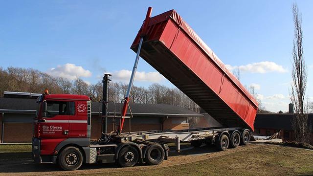 En rød lastbil tømmer en stor container i et åbent område med træer i baggrunden.