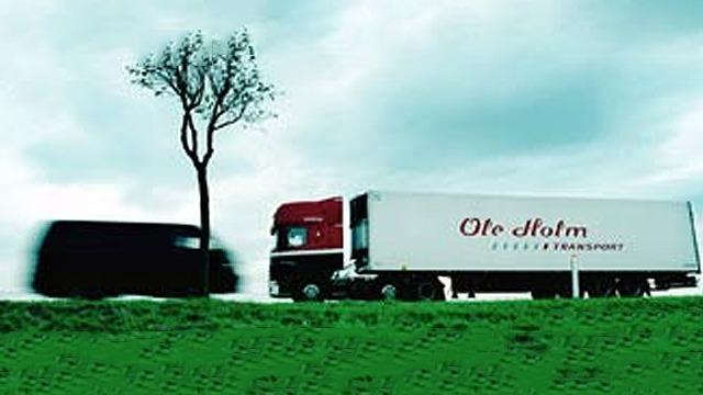 To lastbil fra Ole Holm Transport kører langs en grøn vej under en overskyet himmel.
